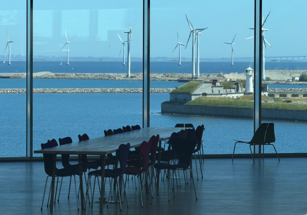 Offshore wind turbines seen through tall windows over a harbour fort and lighthouse